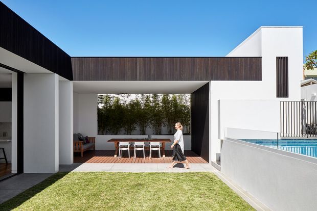Contemporary white-rendered house arranged around a landscaped courtyard, with bold dark timber-clad upper volumes and a connecting bridge-like form. The view shows a covered outdoor dining area, swimming pool edge, lawn and strong geometric facade composition under a clear blue sky.
