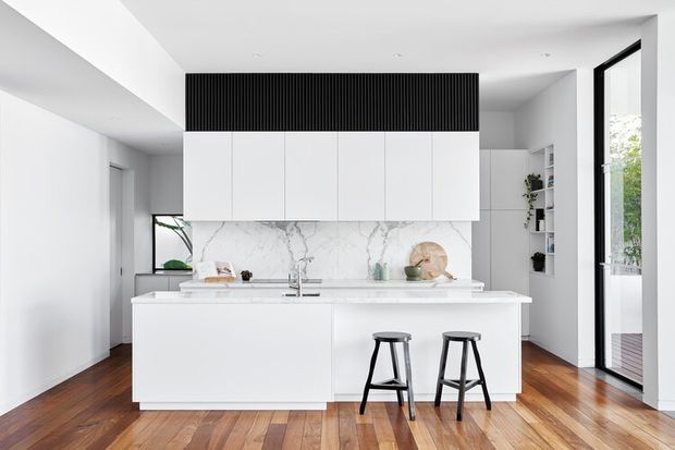 Bright contemporary kitchen with a large white island, two black stools, white flat-panel cabinetry, a marble-look splashback, and a black vertical-ribbed overhead feature. Warm timber flooring contrasts with the predominantly white fit-out, while full-height glazing and open shelving add light and visual openness.