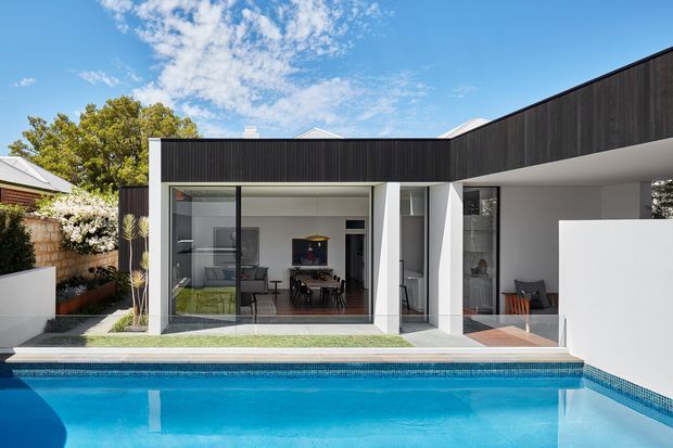 Contemporary single-storey residence viewed from the swimming pool, with a crisp white rendered facade, dark vertical cladding, extensive floor-to-ceiling glazing, and covered outdoor seating. The pool occupies the foreground, while dining and living spaces are visible through the glass. The composition is clean and geometric under a bright blue sky.