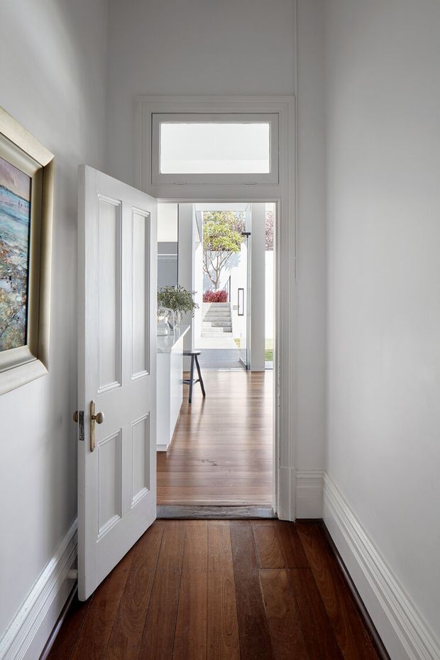 A bright, narrow interior entryway or hallway looking through an open white-panelled door into a larger timber-floored living area and courtyard beyond. The foreground has dark polished timber floorboards, white painted walls, decorative skirting and door trim, a high transom window above the doorway, and framed artwork on the left wall. The space beyond includes light timber flooring, a slim console or stool, contemporary white architectural surfaces, stairs, and planted courtyard elements.