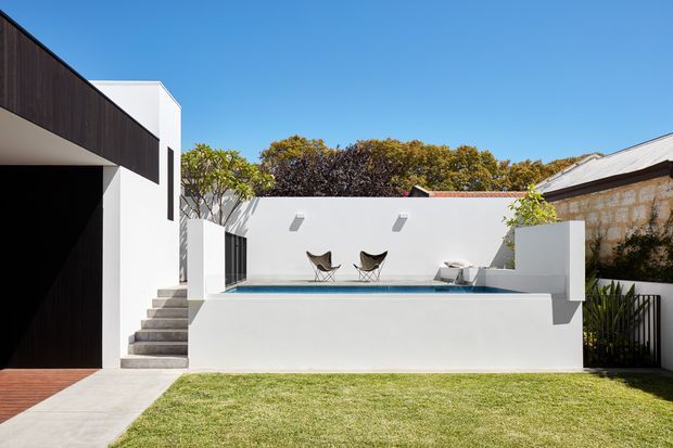 Bright exterior view of a contemporary white-rendered residence with strong geometric forms, dark timber-like cladding, an elevated rectangular swimming pool, integrated steps, and a small paved lawn courtyard. A high boundary wall and adjacent masonry structure enclose the outdoor space under a clear blue sky.