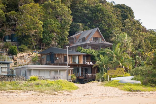 A group of houses set directly beside a sandy beach in a densely vegetated, wooded coastal setting. The main house is a multi-level timber-clad dwelling with a steep gabled roof, dormer-like upper level, balconies, and large glazed openings. A smaller modern timber-and-metal house sits in front, with palms, retaining walls, and a visible cable warning sign contributing to the beachside context.