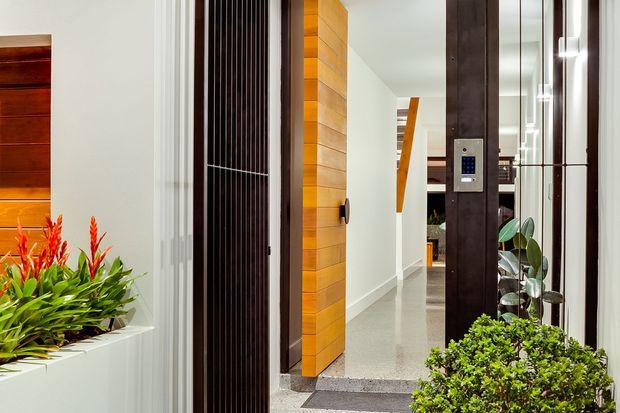 Contemporary residential entry viewed from outside, featuring a timber-clad pivot door, dark vertical security screening, polished concrete entry flooring, white walls, integrated keypad access, and built-in planter beds with tropical greenery. A hallway and staircase are visible beyond the entrance.