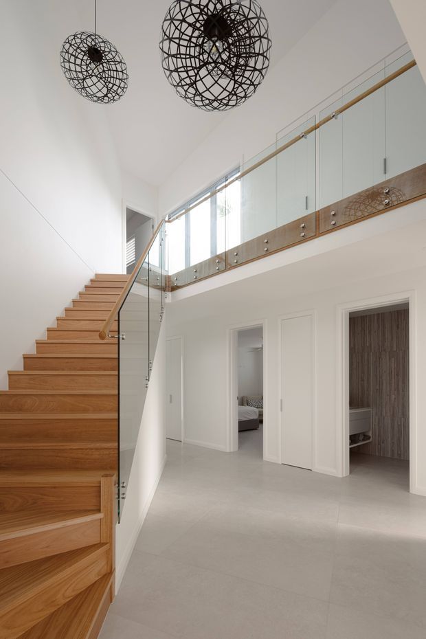 Bright contemporary residential hallway and stairway with a tall open void. A warm timber staircase rises along the left side beside a clear glass balustrade with timber handrail and decorative timber detailing. White paneled walls, pale flooring, upper-level cabinetry, and two sculptural black wire pendant lights create a clean, refined interior. Doorways open to a bedroom and a bathroom or dressing area, while daylight enters through tall glazing.