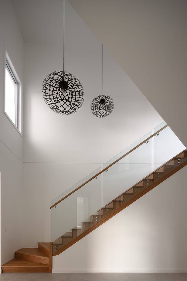 Interior view of a contemporary stairway with warm timber treads and stringers, clear glass balustrades, and slim timber handrails. The stair rises through a tall white-painted void beneath a sloping ceiling, with two suspended black wire-cage pendant lights and a high side window providing natural light.