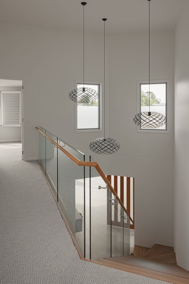 A bright contemporary stairway and upper landing with a carpeted floor, white walls, timber stairs and handrail, and a frameless glass balustrade. Three sculptural black wire pendant lights hang through the stair void, while narrow windows provide natural light. A partially visible bathroom or utility space is seen through the doorway at left.