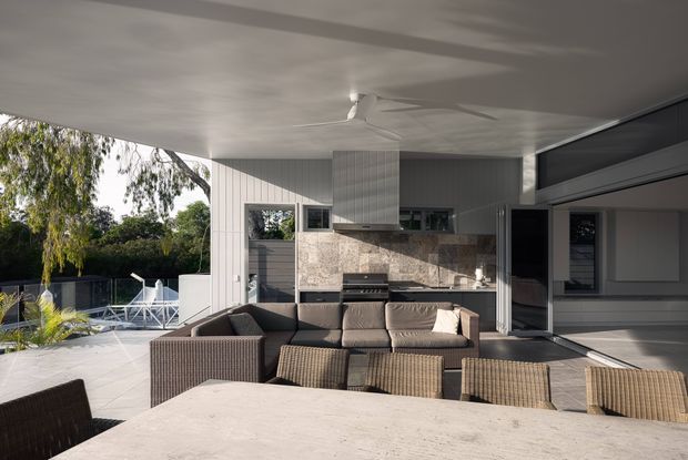 A covered outdoor entertaining area beside an open-plan kitchen, with a large dining table in the foreground, outdoor lounge seating, a built-in barbecue, and a compact kitchen under a broad flat roof. White vertical wall cladding, stone-look splashback, dark-framed glazing, and timber-toned outdoor furniture create a contemporary indoor-outdoor setting. The space opens onto a deck or terrace with garden views.