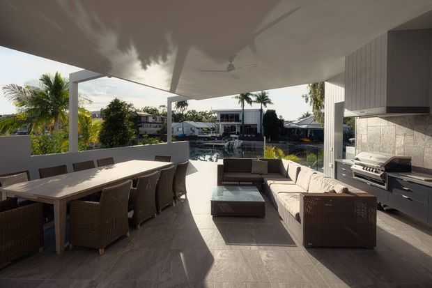 A spacious covered outdoor entertaining terrace with a large dining table, wicker dining chairs, sectional lounge seating, coffee table, and a built-in barbecue kitchen. The terrace overlooks a swimming pool and neighbouring tropical residential setting. The space features a flat roof, pale tiled flooring, white structural columns and vertical wall cladding, with a bright contemporary resort-like atmosphere.