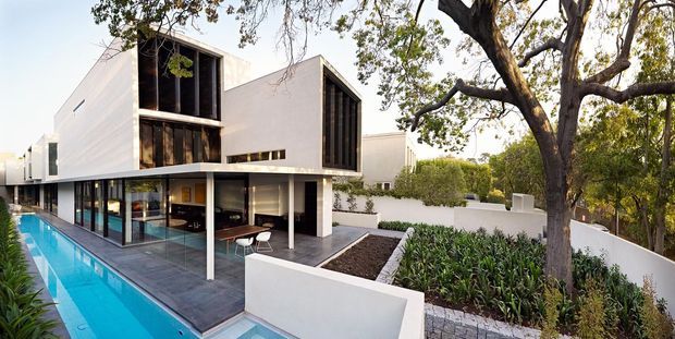 Exterior view of a contemporary multi-level residence with crisp white rendered walls, extensive dark-framed glazing, timber-lined upper openings, and a flat roof. A long narrow swimming pool runs beside the ground floor, while raised landscaped garden beds and mature trees surround the home.