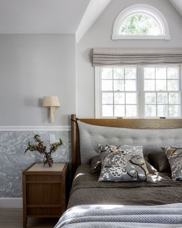 A refined bedroom interior featuring a large upholstered and timber-framed bed, layered grey bedding, patterned decorative cushions, and a dark timber bedside table. Light grey walls are paired with decorative grey lower-wall panelling or wallpaper, white trim, a sculptural wall sconce, and substantial multi-pane windows with an arched transom above. The room has soft natural light and a muted, elegant palette.