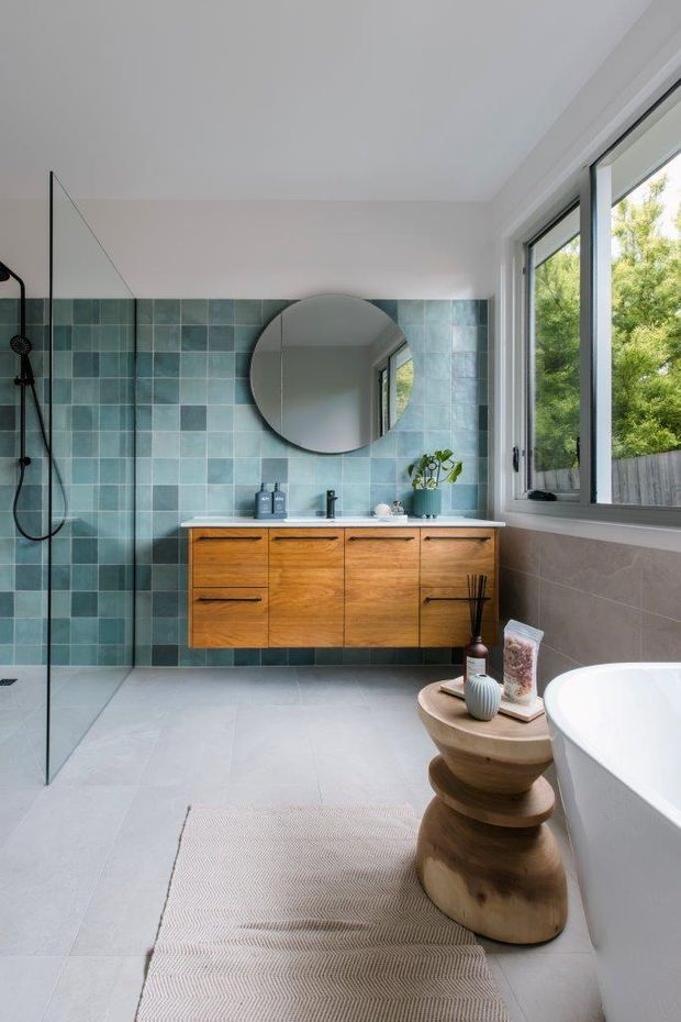 Bright contemporary bathroom with a freestanding white bathtub, frameless glass walk-in shower, wall-mounted warm timber vanity, round mirror, and a blue-green tiled feature wall. Large black-framed windows bring in natural light and overlook greenery. Pale large-format floor tiles and restrained sculptural accessories create a calm, refined atmosphere.