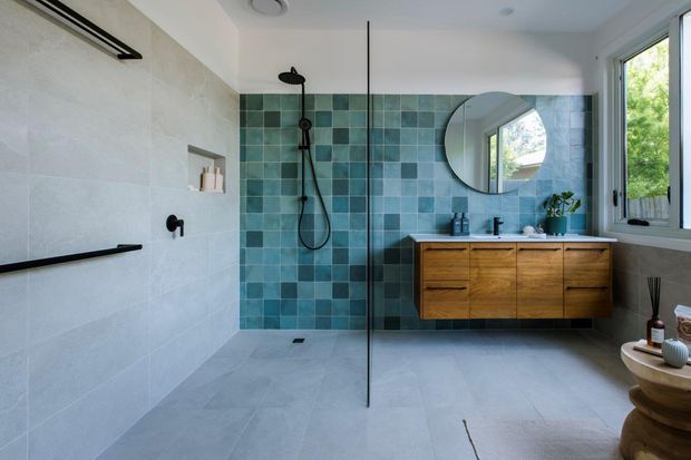 Contemporary bathroom with a spacious walk-in shower enclosed by a frameless glass panel, teal-blue square tiled feature wall, pale large-format wall and floor tiles, and a floating natural timber vanity with a white benchtop and integrated basin. A round mirror, black tapware and shower fittings, recessed niche, skylight, and large window contribute to a bright, refined interior.