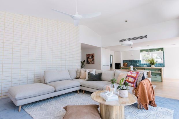 Bright open-plan living room with a large light-grey L-shaped sectional sofa, patterned pale rug, round timber coffee table, soft cushions, and an accent armchair. A compact kitchen with teal cabinetry, tiled splashback, and a window is visible in the background. The room has white walls and ceiling, light timber flooring, abundant daylight, and a ceiling fan.