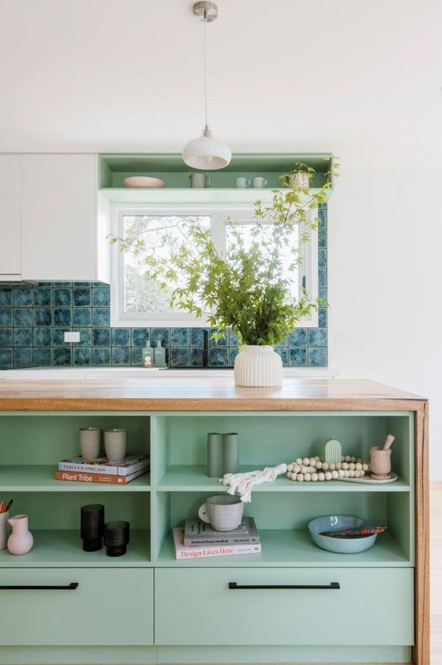Bright, compact kitchen with a pale mint-green built-in island or sideboard featuring open display shelving and lower drawers. The timber benchtop is paired with a glossy blue-green square-tile splashback around a white-framed window. White upper cabinetry, a simple pendant light, and botanical styling create a fresh, playful interior.