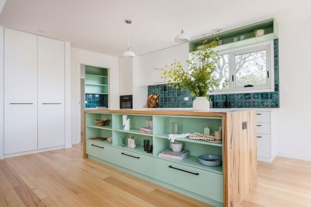 Bright contemporary kitchen with pale timber flooring, white full-height cabinetry, a mint-green island with open shelving and drawers, and a blue-green glazed tile splashback. A timber butcher-block benchtop, integrated appliances, pendant lights, and a large window with greenery visible outside complete the space.
