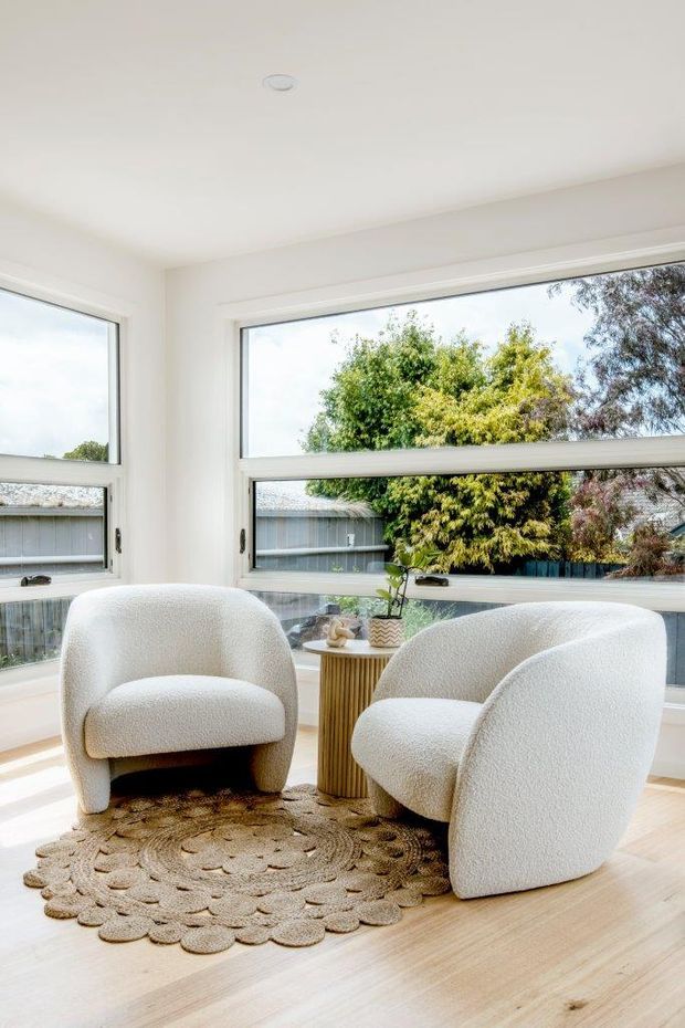 Bright, minimalist sitting area with two rounded boucle armchairs arranged around a small fluted timber side table. The room has white walls and ceiling, pale timber flooring, large white-framed windows, and a neutral textured round rug. Natural light and leafy suburban greenery are visible outside.