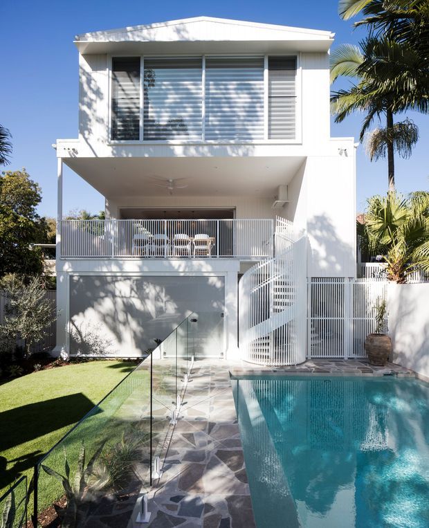 A bright contemporary two-storey white house with a flat roof, expansive louvred upper-level glazing, an elevated covered terrace with white metal balustrades, and an exterior staircase. A swimming pool, paved pool surround, glass safety balustrade, lawn, and tropical palms occupy the foreground and sides.