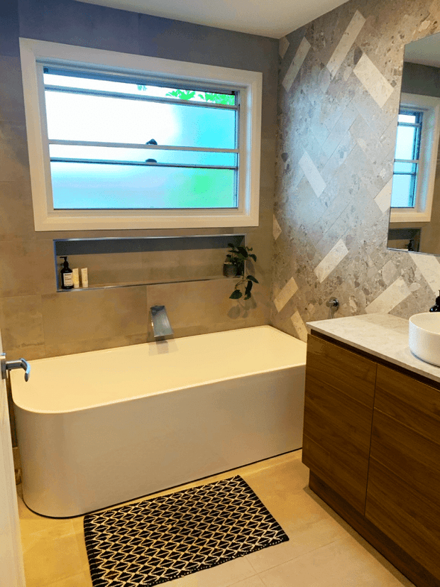 Contemporary bathroom featuring a freestanding white bathtub beneath a frosted horizontal-pane window, a timber vanity with a pale stone benchtop, and a textured geometric feature wall with contrasting light geometric inlays. Warm beige floor and wall tones, recessed shelf, mirror, and black patterned bath mat complete the space.