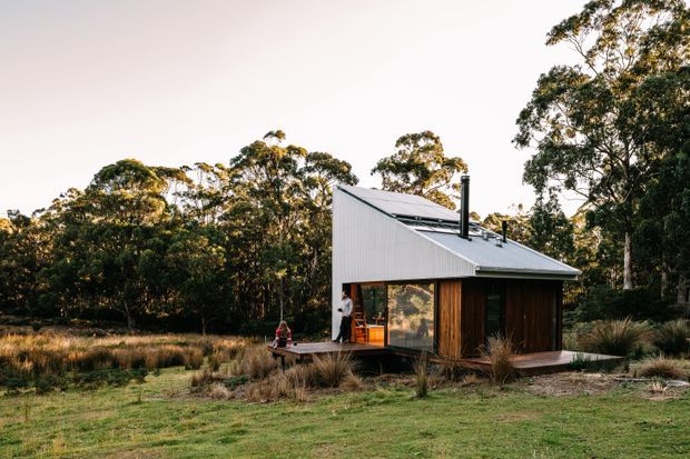 A compact contemporary cabin set within a natural bushland landscape. The small single-storey structure has a steep asymmetrical gabled metal roof, vertical white corrugated cladding, dark timber wall cladding, large glazed openings, and a small timber deck. Native trees and grasses surround the building.