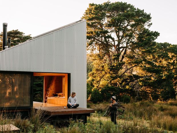 Exterior view of a compact contemporary cabin set within a natural bushland setting. The building has a sharply pitched, asymmetrical roofline, pale vertical corrugated cladding, dark timber wall panels, and a warm timber-lined entry or living opening onto a raised timber deck. Two people sit and stand near the deck, surrounded by mature trees and tall grasses.