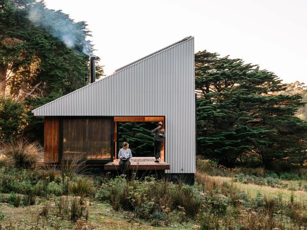 A compact contemporary cabin set within a dense natural forest landscape. The building has a sharply sloping single-pitch roof clad in vertically ribbed light-grey metal, dark timber wall cladding, large glazed openings, and a small timber deck with two people. Native grasses and mature trees surround the structure, and smoke rises from a slim roof chimney.