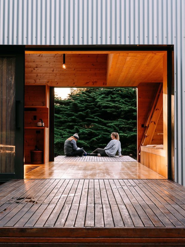 A small timber-lined retreat or cabin with a large open front connecting the interior to a raised timber deck. Warm wood walls, ceiling, cabinetry and flooring frame a view of dense evergreen vegetation, with two people seated at the threshold. Corrugated light-coloured metal cladding surrounds the opening, and the scene has a warm, tranquil indoor-outdoor character.