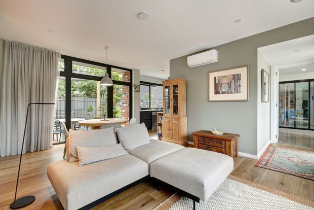 A bright, contemporary living room with a large light-grey sectional sofa, timber flooring, and neutral grey walls. Full-height curtains frame black-framed glazed doors opening to an outdoor area, while a timber display cabinet, sideboard, framed artwork, pendant light, and patterned rug add warmth and character. A hallway connects to additional rooms in the background.