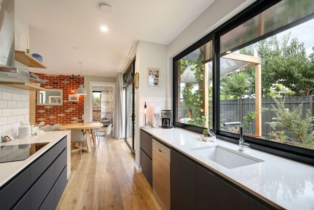 A bright contemporary kitchen and dining area with black lower cabinetry, white tiled splashback, white stone benchtops, timber flooring, and large black-framed windows overlooking a landscaped backyard. A brick feature wall and compact dining table add warmth, while a long galley-style layout connects the kitchen to the dining space.