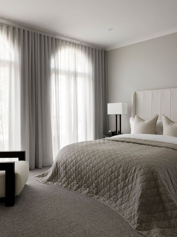 A softly lit, refined bedroom in a neutral palette. A large upholstered bed with a tall textured headboard is dressed in a quilted taupe-grey bedspread and layered pillows. Floor-to-ceiling sheer and grey curtains frame a bright window, while a simple bedside lamp and dark side table provide contrast. The room has carpeted flooring, light grey walls, and restrained contemporary styling.