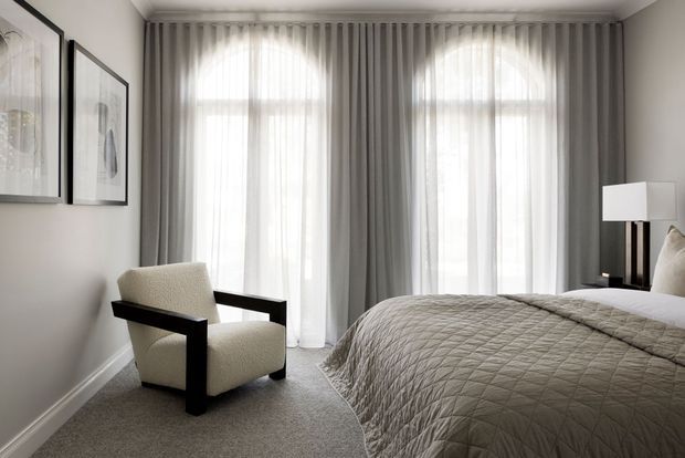 A serene, refined bedroom with a neutral palette, tall arched windows dressed in full-height sheer and grey curtains, a quilted grey bedspread, an upholstered black-framed armchair, framed artwork, and a simple bedside lamp. The room has soft natural light, carpeted flooring, and classic wall and ceiling detailing.