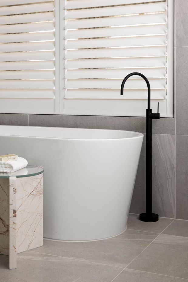 A contemporary bathroom featuring a freestanding white bathtub, matte black floor-mounted bath tap, grey tiled walls and floor, and white plantation shutters over the window. A small marble-look side table with folded towels sits beside the tub.