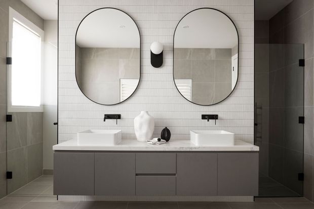 Contemporary double-vanity bathroom with a wide floating grey vanity, two white vessel basins, black wall-mounted tapware, and paired rounded-arch mirrors. Large-format grey wall and floor tiles are contrasted with a small white grid-tile feature wall, while a glass shower enclosure is visible on the right.