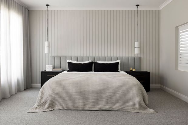 A serene, contemporary bedroom with a neatly made bed, upholstered vertical-panel headboard, dark bedside tables, pendant lights, sheer curtains, plantation shutters, and soft grey carpeting. The palette is neutral and understated with balanced natural light.