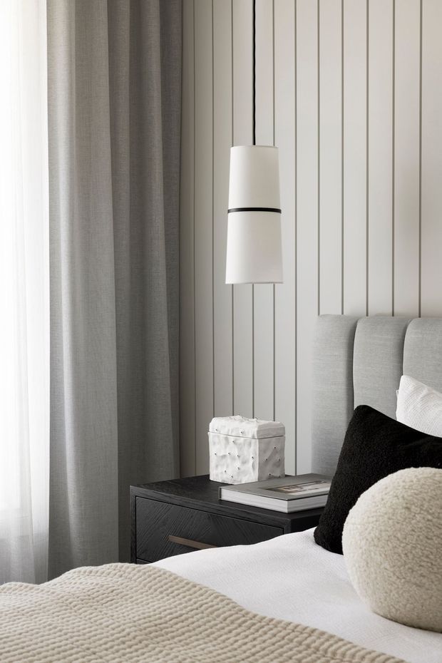Close-up of a softly styled bedroom corner with a tall upholstered grey headboard, layered white and neutral bedding, black and cream accent cushions, and a dark timber bedside table. A simple white pendant light hangs in front of a vertical grey wall-panelled feature wall, with full-height grey curtains beside the window. The overall look is restrained, monochromatic, and contemporary.