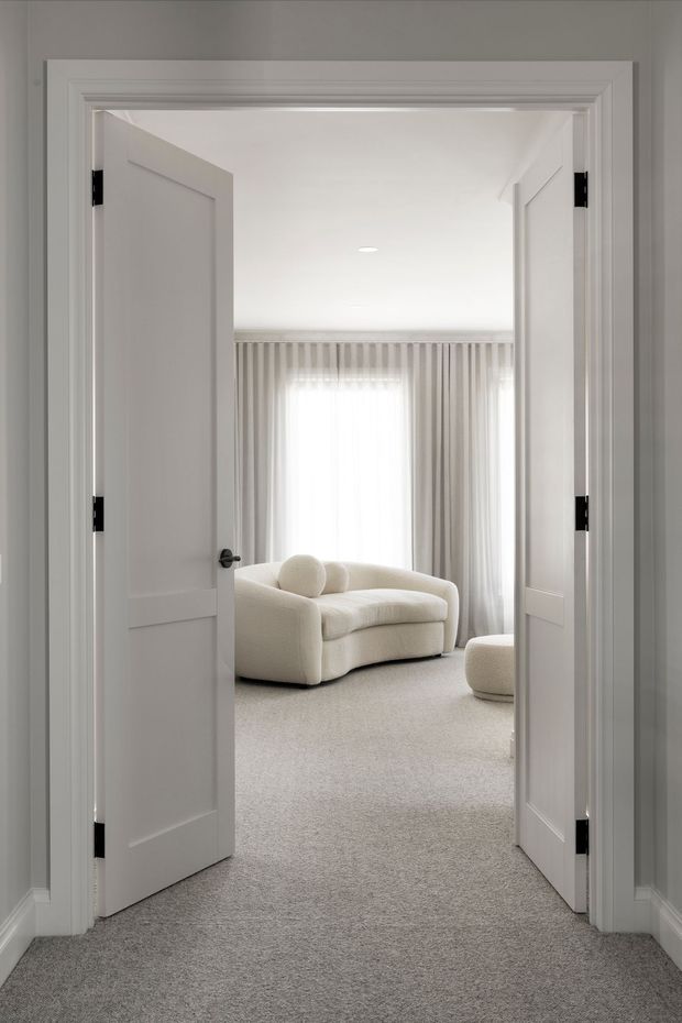 A bright, monochromatic interior view through open double doors into a softly furnished living room. The space features white paneled doors and decorative trim, pale grey carpet, sheer floor-to-ceiling curtains, and a rounded cream upholstered sofa with a matching ottoman. The room has a calm, refined, minimalist appearance.