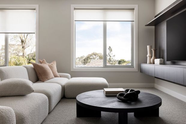 Contemporary living room with a large curved modular sectional sofa, round black coffee table, neutral carpet, and a dark built-in media wall with floating cabinetry and open shelving. Large windows with roller blinds provide soft natural light and views of trees and neighbouring rooftops.