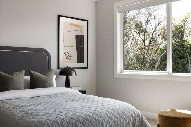 A bright, softly styled bedroom featuring a neatly made bed with a dark upholstered headboard, textured grey bedding, muted green cushions, a black dome table lamp, and framed abstract artwork. A large white-framed window brings in natural light and overlooks greenery. The palette is neutral and calm with understated contemporary furnishings.