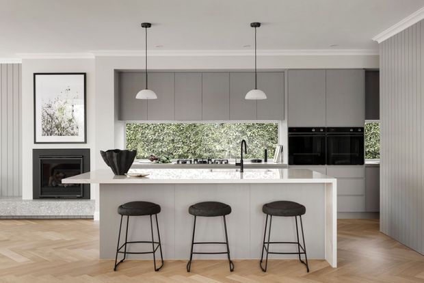 Bright contemporary kitchen with a large white waterfall-edge island, three black bar stools, grey full-height cabinetry, integrated black appliances, and a wide window overlooking greenery. The room features light herringbone timber flooring, vertical wall panelling, pendant lights, a fireplace, and refined white moulded ceiling trim.