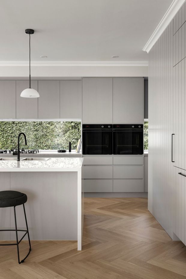 A bright, refined contemporary kitchen with soft grey full-height cabinetry, integrated double ovens, a white stone island with sink and black tapware, and a black pendant light. The kitchen opens to leafy greenery through a large window, while natural timber herringbone flooring and vertical wall panelling add warmth and texture.