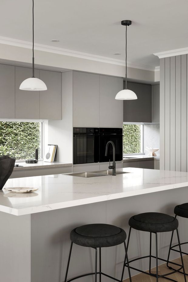 A refined contemporary kitchen in soft grey and white tones, featuring full-height flat-panel cabinetry, integrated black appliances, a large white stone island with undermount sink and dark curved tap, and three black upholstered bar stools. Pendant lights hang above the island, while vertical wall panelling and generous windows add architectural detail and natural light.