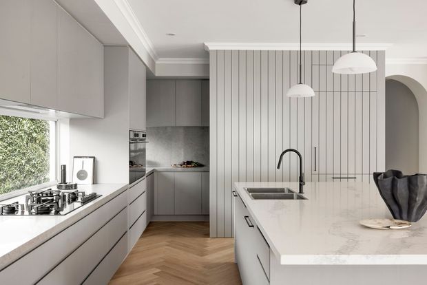 A refined contemporary kitchen with a large white stone island, integrated sink and black tapware, streamlined grey cabinetry, a gas cooktop beneath a window, and a vertical fluted feature wall concealing the rear kitchen area. Herringbone timber flooring, arched wall opening, pendant lights, and crisp white architectural detailing create a bright, polished interior.