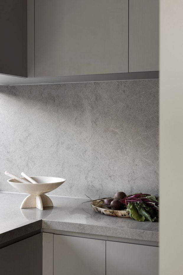 Close-up view of a refined contemporary kitchen corner with matte grey cabinetry, a continuous grey stone benchtop and matching stone splashback. A sculptural ceramic bowl and a tray of vegetables sit on the counter, while under-cabinet lighting softly highlights the textured stone surface.