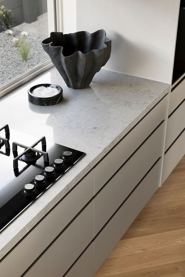 Close-up view of a refined contemporary kitchen featuring a pale stone countertop, integrated light cabinetry with slim dark reveals, and a black gas cooktop. A sculptural black vase and small bowl sit beside a large window overlooking a minimalist gravel garden. Warm timber flooring adds contrast.