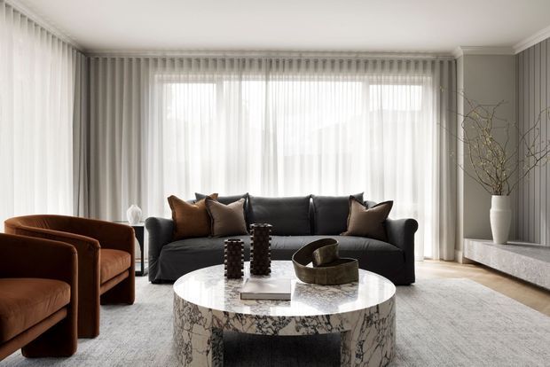 Elegant contemporary living room with full-height sheer curtains, a dark charcoal sofa, rust-brown upholstered armchairs, a round white marble coffee table, light carpet, and a minimalist stone media console. Neutral tones and soft daylight create a refined, serene atmosphere.