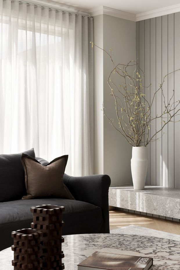 A refined contemporary living room with a dark upholstered sofa, taupe cushions, sheer full-height curtains, grey painted walls, decorative vertical wall panelling, and a pale stone floating media cabinet. A tall white vase with bare branches adds an organic feature, while a veined marble coffee table and sculptural dark timber decor complete the elegant neutral scheme.