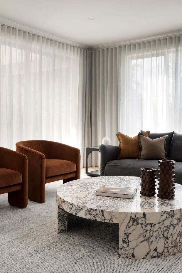 A refined living room with full-height sheer curtains, a dark charcoal sofa, two deep rust-brown armchairs, and a large round marble coffee table with dramatic veining. The room has a soft neutral palette, plush grey carpet, restrained decorative objects, and abundant filtered daylight.