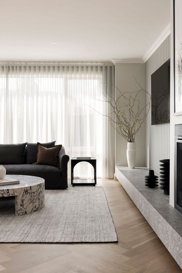 Refined contemporary living room with a black sofa, sculptural marble coffee table, pale herringbone timber flooring, large sheer-curtained glazing, and a textured media wall with vertical panelling, television, fireplace, and low stone console. Neutral tones and restrained black accents create a calm, luxurious atmosphere.