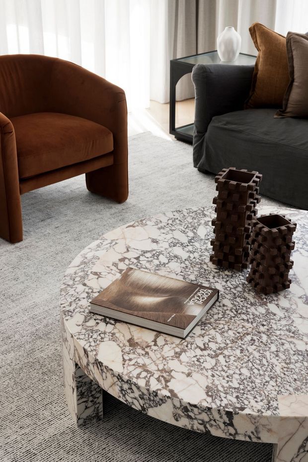 A refined living room vignette with a rounded rust-brown upholstered armchair, a dark charcoal sofa with warm brown cushions, sheer floor-to-ceiling curtains, and a black-framed side table. A large round coffee table clad in heavily veined white-and-grey stone dominates the foreground, styled with a book and sculptural dark timber objects. The space has a soft, warm, contemporary feel with layered neutral textures.