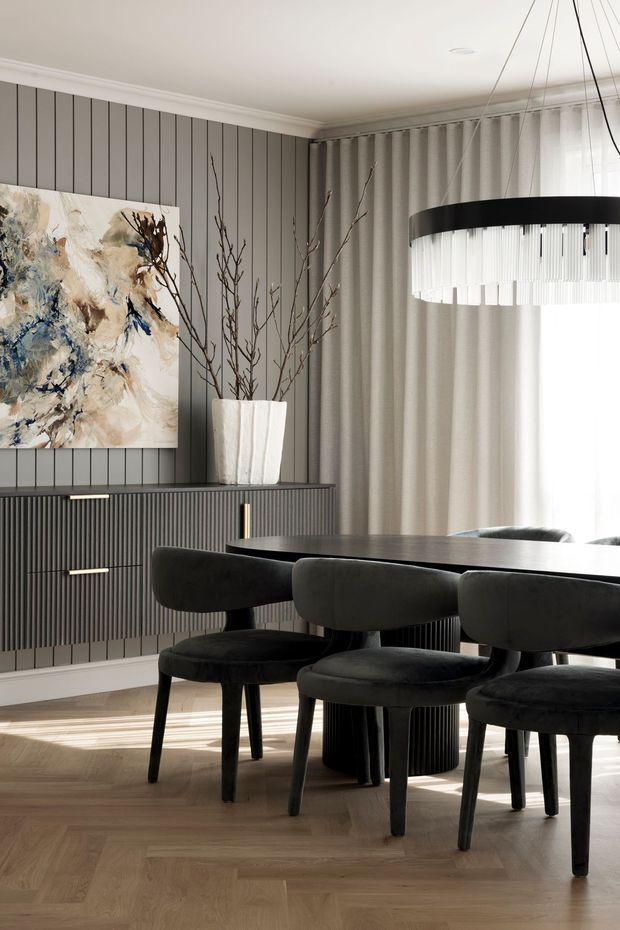 A refined dining room with a dark oval dining table and upholstered black dining chairs, set against grey vertical-panelled walls. A ribbed dark sideboard with brass pulls, a large abstract artwork, sculptural branch arrangement, layered curtains, and a statement circular pendant light create a sophisticated, softly lit interior.