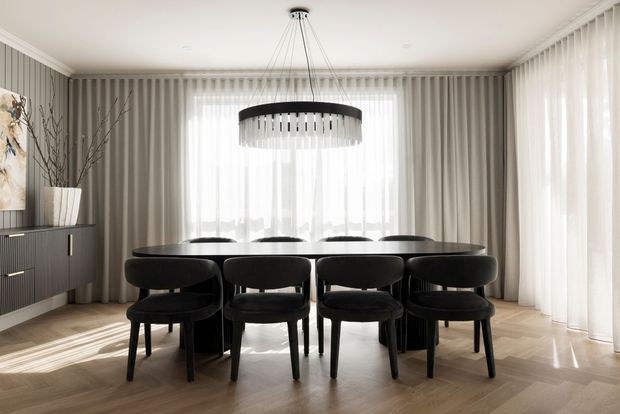 A refined dining room with a large round dark dining table and matching black upholstered chairs, positioned in front of full-height sheer curtains and bright windows. The room features light timber flooring, grey vertical wall panelling, a dark sideboard, and a contemporary circular pendant chandelier.